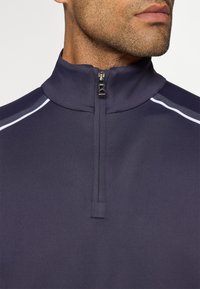 Pull en navy avec un col montant, fermeture éclair quart et des lignes blanches aux épaules. Finition en tissu lisse et texturé.