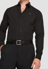 Chemise noire à manches longues avec un col à boutons et un petit logo blanc sur la poitrine, associée à une ceinture noire avec une boucle argentée.