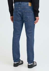 Blauwe spijkerbroek met een rechte snit, vijfzakkenontwerp, zichtbare stiksels en een tan stoffen label op de tailleband, gecombineerd met zwarte sneakers.