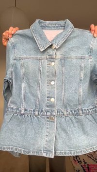 Lyseblå denimjakke med sølvknapper, strukturerede sømme og en indsnævret talje, holdt op af to hænder mod en neutral baggrund.