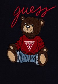 Bestickter Teddybär, der einen roten Pullover mit einem Dreieck-Logo und blaue Jeans trägt, vor einem marineblauen Hintergrund, mit detaillierten Felltexturen.
