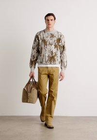 Brown and gray patterned sweatshirt, beige pants, and a tan bag with dark accents. Model walking on a neutral background.
