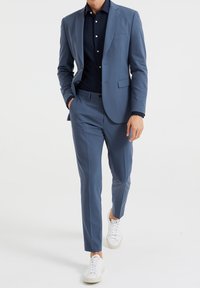Ljusblå kostym med skräddarsydd kavaj och smala byxor, kombinerad med en marinblå skjorta med knappar och vita sneakers. Slätt tyg, minimal detaljering.