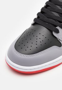 Graue und schwarze Sneaker mit einem glatten Lederoberteil, perforierter Zehenpartie für Atmungsaktivität, schwarzen Schnürsenkeln und roter Gummisohle. Weiße Nähten.