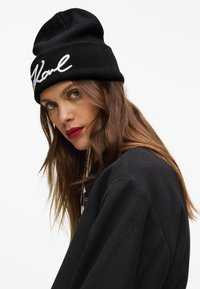 Žena s dlouhými hnědými vlasy, která m�á na sobě černou čepici s bílým nápisem a černou bundu, se dívá mírně přes rameno na bílém pozadí.