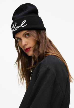 Woman with long brown hair wearing a black beanie with white script and a black jacket, looking slightly over her shoulder against a white background.