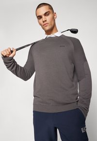 Grå strikket genser med ribbestrikkede detaljer og rund hals, med en logo på brystet, båret av en modell med en golfklubb.
