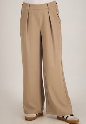 Beige broek met hoge taille en wijde pijpen, met plooien aan de voorkant, zijzakken en riemlussen, gecombineerd met witte sneakers met zwarte strepen.