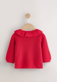 Rdeč pleten pulover z vzorcem teksture, z okroglim, nabranskim ovratnikom in dolgimi rokavi. Visí na lesenem obešalniku na svetli podlagi.