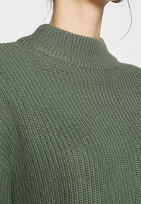 Zielony dziany sweter z wysokim, prążkowanym kołnierzem i teksturowanym materiałem. Projekt cechuje luźny krój z subtelnym połyskiem.