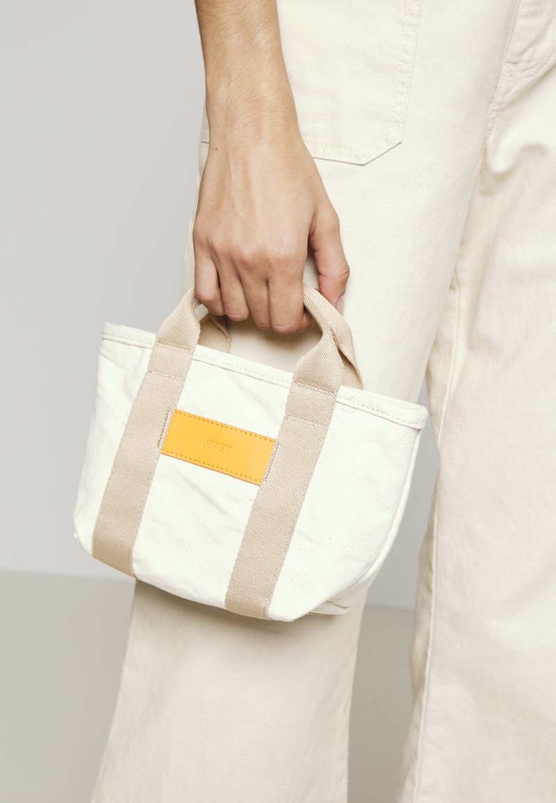 Main tenant un petit sac en toile écrue avec des bretelles beiges et un patch rectangulaire jaune, portant un pantalon écru.