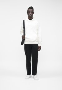 Sudadera blanca con capucha, pantalones negros de corte holgado y zapatillas blancas. El modelo lleva un bolso negro al hombro, posando sobre un fondo liso.