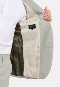 Man in light gray suit jacket showing beige and dark brown lining with a black "JEFF" label inside.
