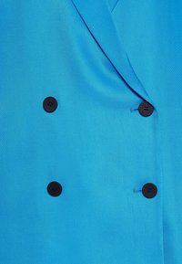 Niebieska, teksturowana marynarka wykonana z tkaniny, charakteryzująca się dwurzędowym krojem z trzema czarnymi guzikami i spiczastymi klapami.