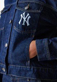 Denim jakna tamnoplave boje sa srebrnim detaljima. Ima dva džepa na grudima, kontrastne šavove i bijeli izvezeni logo na prsima.