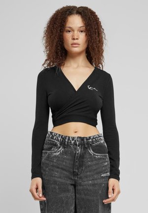 Schwarzes Langarm-Wickeltop mit V-Ausschnitt, versehen mit einem kleinen weißen Logo. Kombiniert mit hoch taillierten, verwaschenen grauen Jeans.