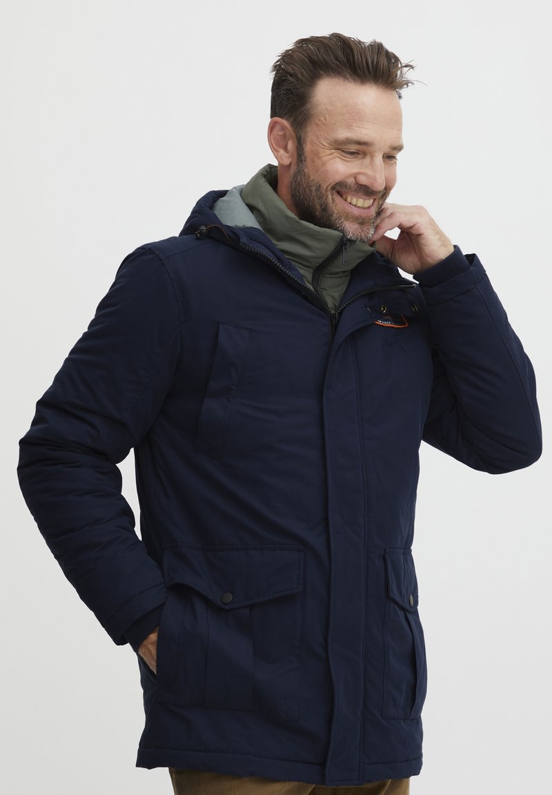 Navy blue insulated jacket with adjustable hood, front pockets, and a zip closure. Features a gray inner layer for added warmth.