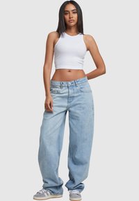 Young woman wearing white sleeveless crop top, baggy light blue jeans, and gray sneakers, standing with one hand behind her back.