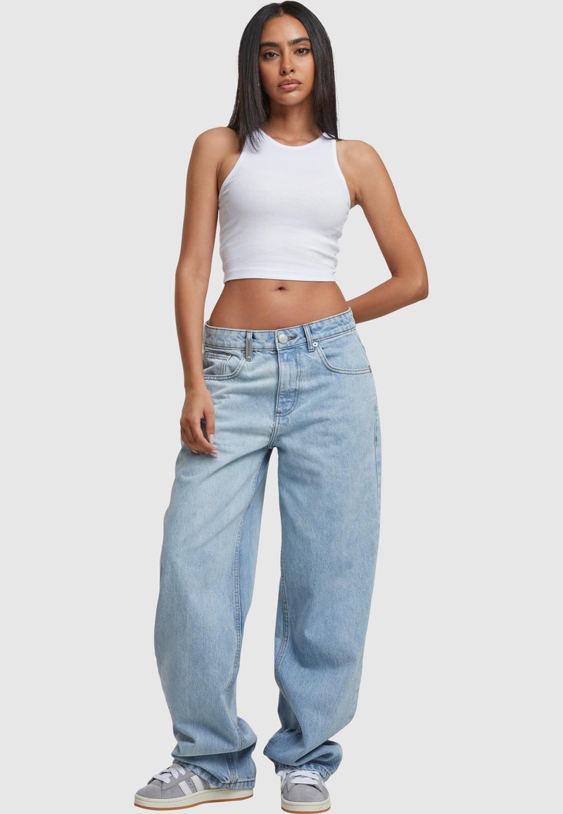 Young woman wearing white sleeveless crop top, baggy light blue jeans, and gray sneakers, standing with one hand behind her back.