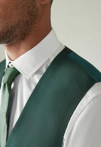 Green textured vest with a white dress shirt and a matching green tie. The vest features a smooth fabric on the shoulders for contrast.
