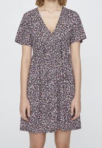 Donna che indossa un vestito nero a maniche corte e scollo a V con un piccolo motivo floreale rosa e bianco, in piedi contro uno sfondo semplice.