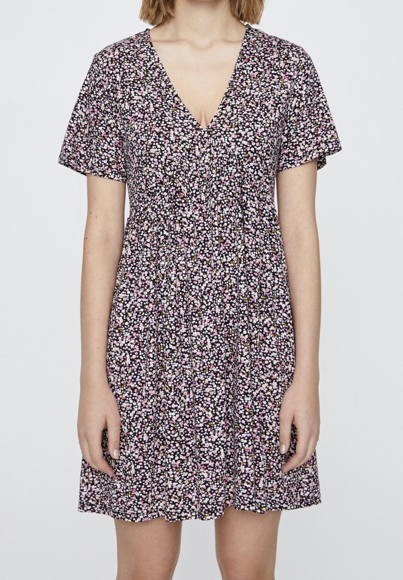 Donna che indossa un vestito nero a maniche corte e scollo a V con un piccolo motivo floreale rosa e bianco, in piedi contro uno sfondo semplice.