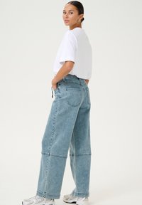 Ljusblå denim byxor med vida ben och avslappnad passform samt hög midja. Har framfickor och en sömdetalj nära fållen. Kombineras med en vit t-shirt.