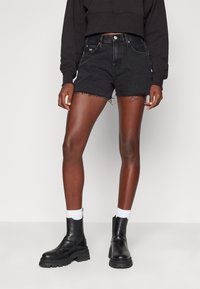 Person med svarta slitna denimshorts, svart kort sweatshirt, vita strumpor och svarta chunky ankelskor, som står mot en enkel bakgrund.