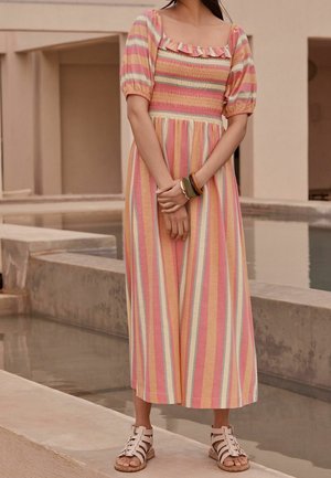 Kvinde iført en pastelfarvet stribet midi-kjole med puffede ærmer og flæsekrave, stående ved et betonkildeanlæg, iført hvide sandaler.