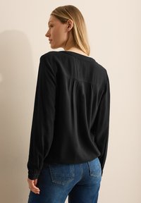 Zwarte blouse met lange mouwen van een textuurstof en geplooid achterdetail, gecombineerd met blauwe jeans, weergegeven vanuit een zijaanzicht.