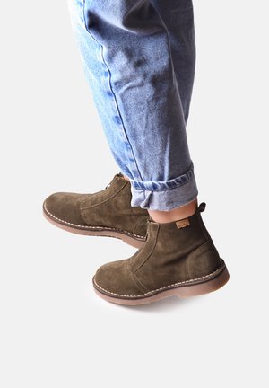 Botas de tobillo de ante verde oscuro con punta redonda, suela de goma marrón y detalles de costura. Combinadas con unos vaqueros de mezclilla azul claro remangados.