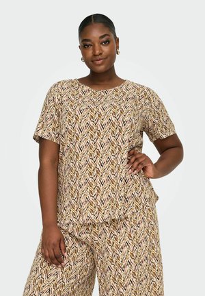 Kvinne med mørk hud og trukket hår, iført matchende beige mønstret kortermet topp og vide bukser, poserer selvsikkert med hånden på hoften.