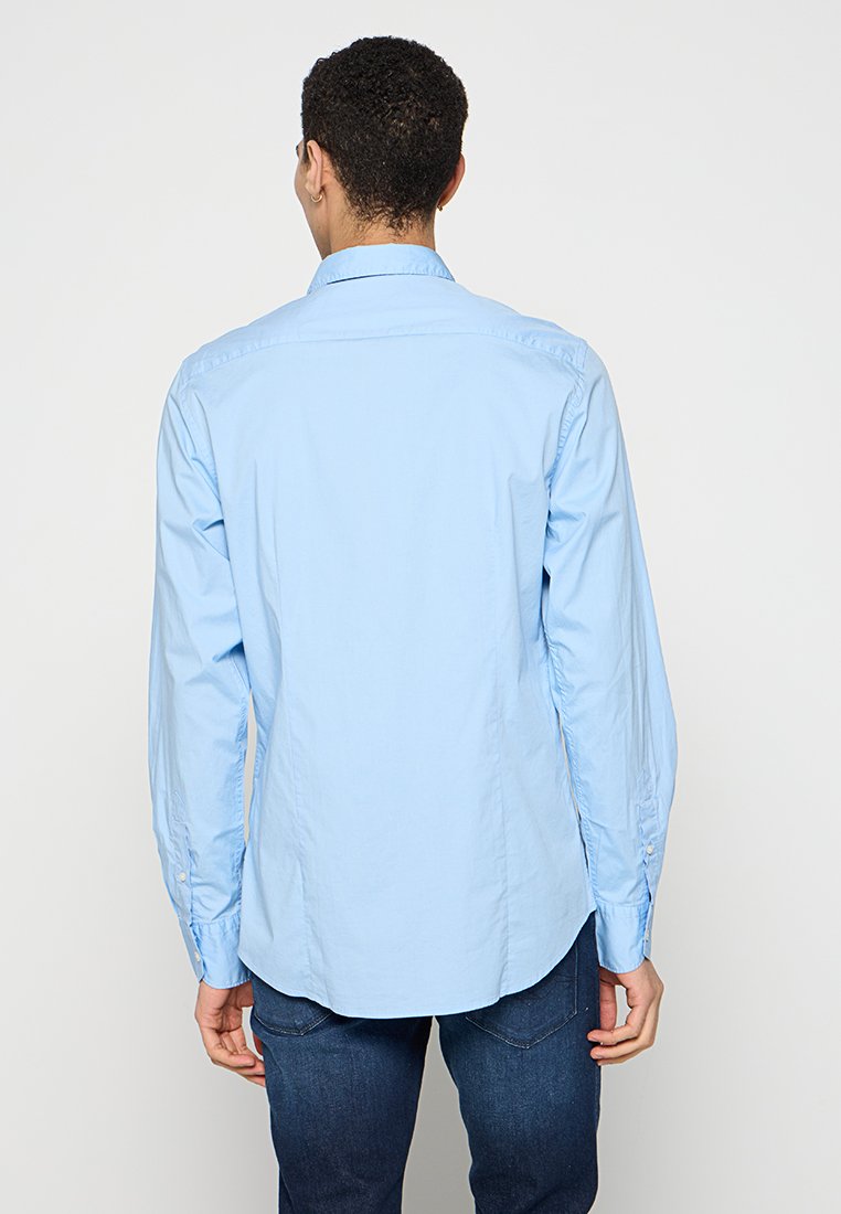 Camicetta azzurro chiaro a maniche lunghe in cotone, dotata di colletto button-down, polsini con bottoni e orlo curvo. Indossata con jeans scuri.