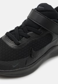 Chaussure de sport noire avec une tige en mesh, des accents noirs lisses, un collier rembourré et une sangle de maintien sécurisée au niveau du cou-de-pied pour un soutien optimal.