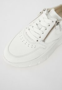 White leather sneaker with textured finish, featuring a lace-up front, perforated toe, and side zipper accent. Chunky white sole.