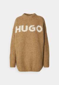 Pull en maille avec une texture côtelée de couleur marron clair, un large col rond et une coupe surdimensionnée. "HUGO" est affiché en blanc de manière bien visible.