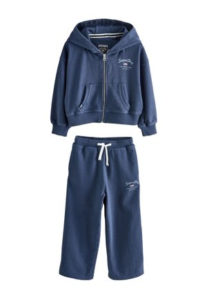 Mädchen trägt einen marineblauen Kapuzenpullover mit Reißverschluss über einem Jeanshemd, passende marineblaue Hose und beige Slip-On-Schuhe, steht mit einer Hand am Kopf.