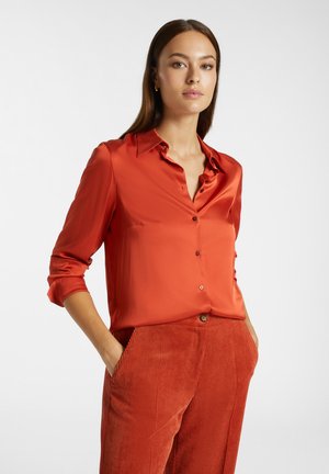 Camicia rossa di raso con bottoni, colletto e maniche lunghe, abbinata a pantaloni in velluto a coste dello stesso colore. Texture liscia, design strutturato.