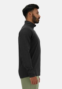 Chemise noire à manches longues avec un col haut, présentant une texture lisse et un design ajusté, portée par une personne de profil sur un fond uni.