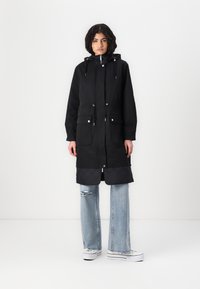 Lang svart parka med quiltet hem, justerbar hette, frontlommer og metalliske detaljer. Kombinert med lyseblå vide jeans og hvite sneakers.