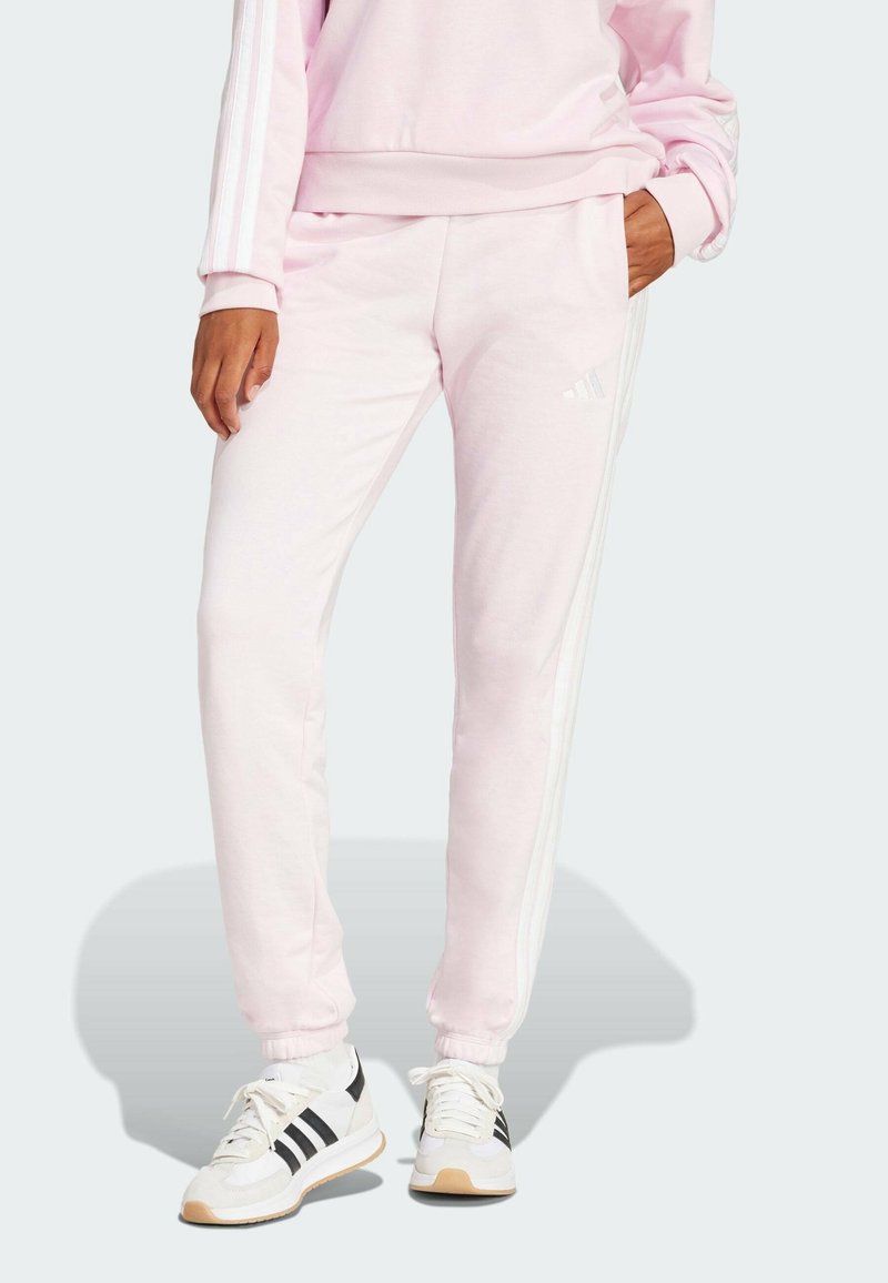 Pantaloni della tuta rosa chiaro realizzati in tessuto morbido, con accenti bianchi a tre strisce lungo i lati e una vestibilità rilassata alle caviglie.