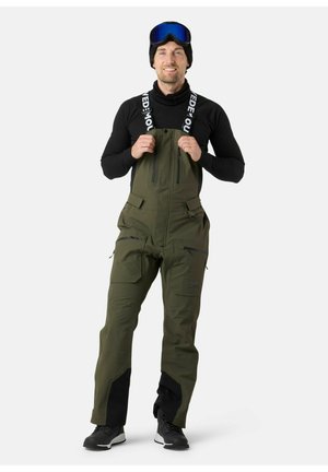 Pantaloni salopette impermeabili verdi con bretelle regolabili, zip frontale, numerose tasche con zip e dettagli rinforzati neri ai polsini.