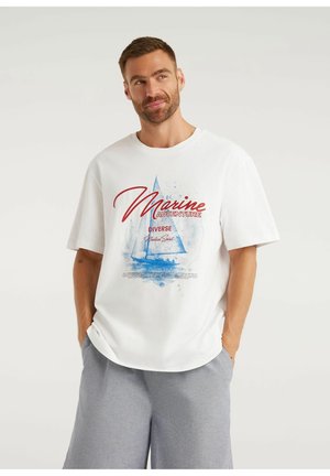 Mężczyzna w białej koszulce z niebieskim żaglowcem i czerwonym napisem "Marine Adventure", w połączeniu z jasnoszarymi szortami, ręce w kieszeniach, patrzy w górę.