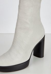 Stivale ankle bianco in pelle con una texture liscia, punta rotonda e tacco spesso nero, caratterizzato da un design minimalista e un leggero lucido.