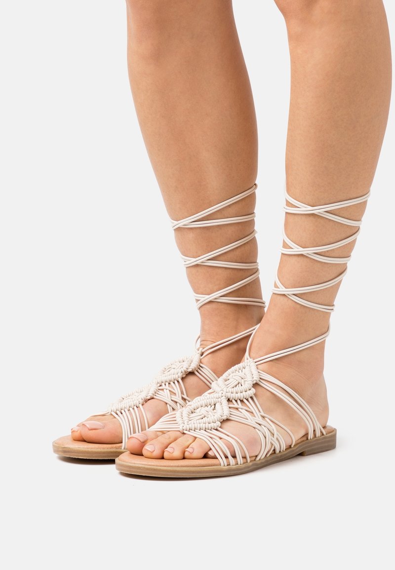 Piedi che indossano sandali piatti beige con intricate cinghie intrecciate che avvolgono caviglie e polpacci su uno sfondo semplice.