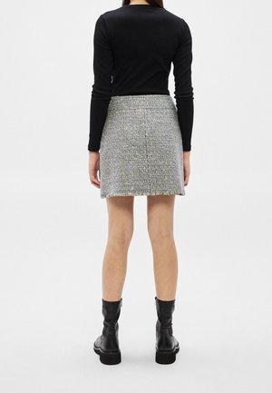 Vrouw die een zwart top met lange mouwen, een grijze mini rok van tweed en zwarte enkellaarsjes draagt, staand met haar rug naar de camera op een eenvoudige witte achtergrond.