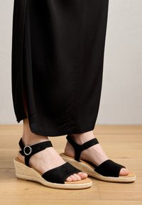 Sandales compensées en suédine noire avec une semelle en espadrille, présentant une sangle de cheville ajustable et un accent métallique en forme de cercle.