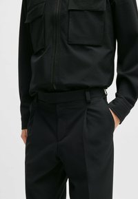 Schwarzes, strukturiertes Stoffoutfit mit einer Jacke mit Reißverschluss und zwei Brusttaschen sowie einer maßgeschneiderten Hose mit einem glatten Gürtel.