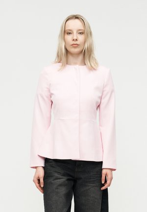 Γυναίκα με ξανθά μαλλιά που φοράει ένα ροζ ανοιχτόχρωμο στενό σακάκι και μαύρο τζιν, στέκεται μπροστά σε ένα απλό λευκό φόντο.