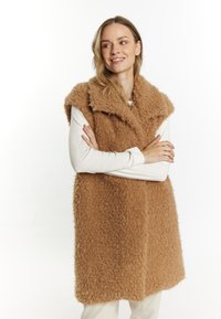 Bruine pluizige mouwloze vest met een sjawlboord en textielstructuur, met een rechte snit en zacht afwerking. G gedragen over een witte longsleeve.